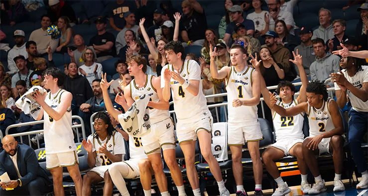UC San Diego men's basketball team members cheer and throw signs from the LionTree Arena bench
