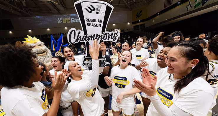 UC San Diego women's basketball team members cheer and laugh while holding and gathering around the Big West trophy