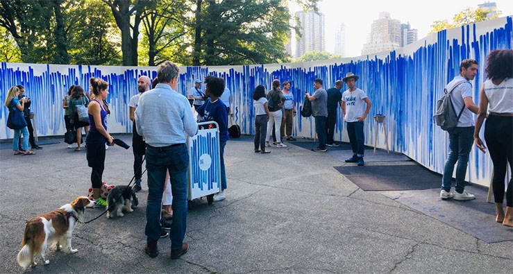 A group of people stand outdoors near a large, long continual canvas decorated with vertical blue paint strokes - Photo by Jan Strempel / Studio Jeppe Hein