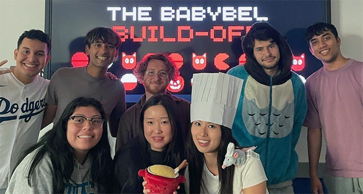 A group of 8 smiling students pose for the camera - one wears a chef hat and holds a small bowl of cheese dip
