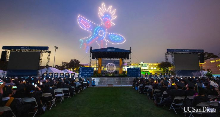 A large stage with many rows of cap-and-gown-clad students in front of it -- it's dusk and a multicolored drone show is displaying in the sky above the stage -- it's a little Sun God ;)