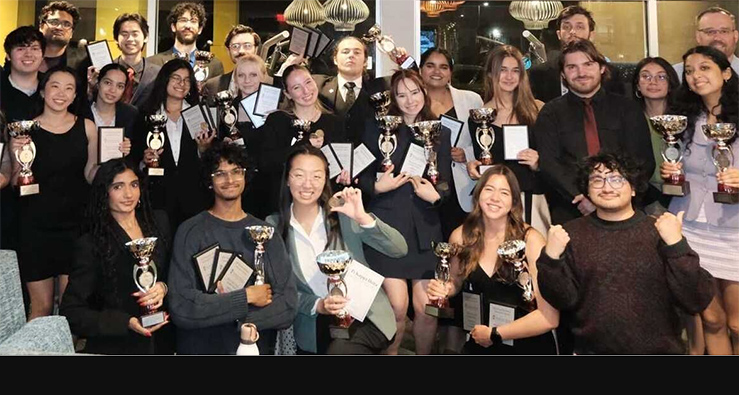 Large group of smiling, happy debate-team students hold up golden trophies