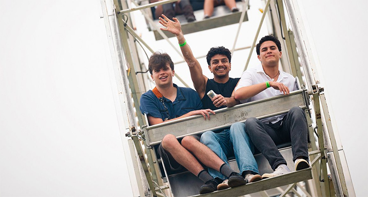 Three students sit on a Ferris wheel cart going - one is waving