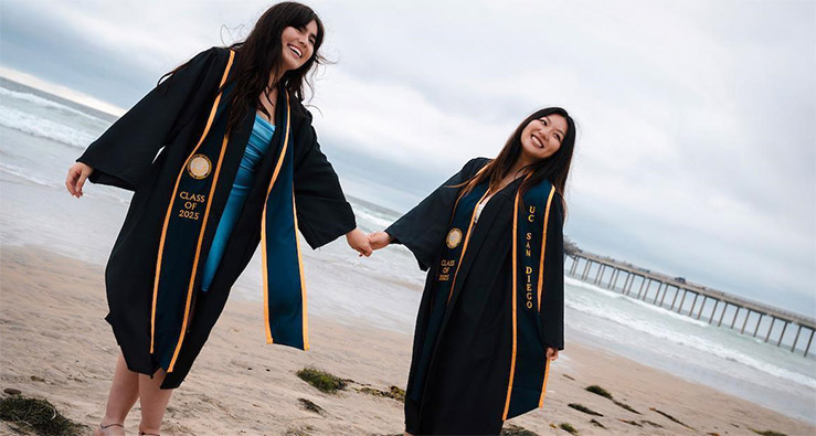 Two smiling UCSD graduates in their caps and gowns and stoles, posing on the beach near campus