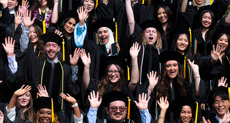 A large group of pharmacy students in black mortarboard caps and gowns smile, laugh, and lift up their hands in the air