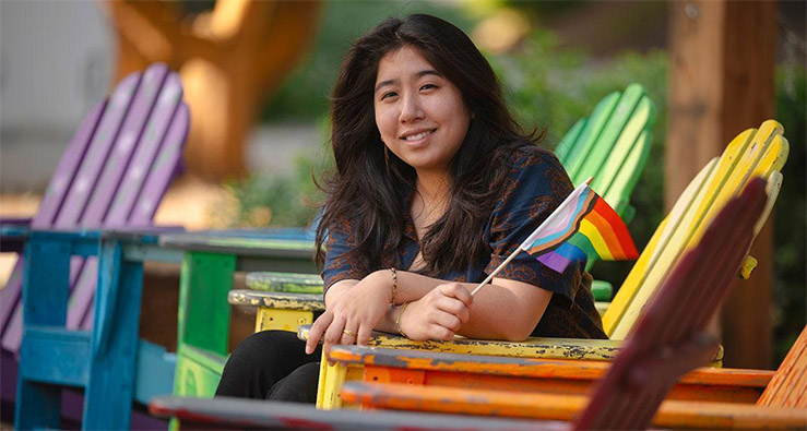 A dark-haired woman sits and smiles, holding a rainbow colored Pride flag, surrounded by other rainbow-colored decorations