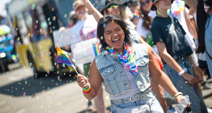 A smiling person waves a small rainbow flag as they march down the street, with colorful confetti all around