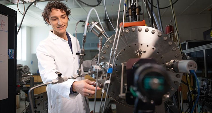 UC San Diego Qualcomm Institute Research Specialist Riley Need wears a white lab coat and stands before a large metal machine with many wires coming into it from all directions, tinkering with a dial