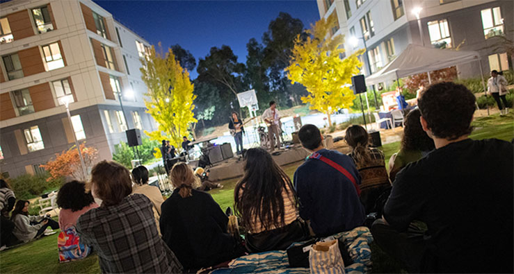Students sit on the Sixth College lawn at night, watching a musical performance