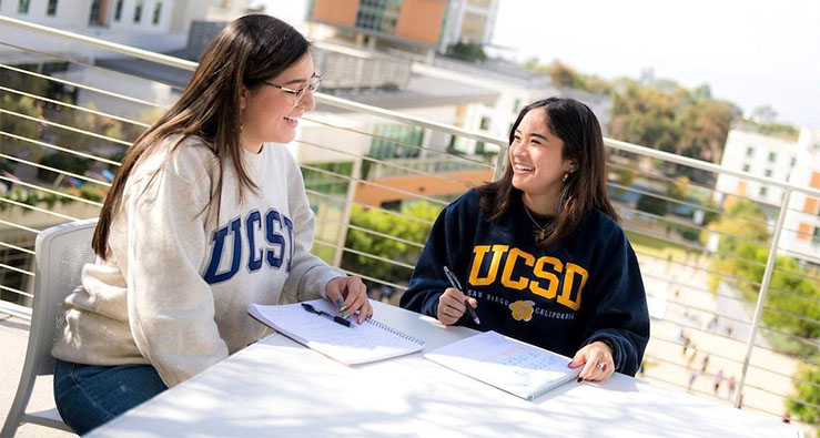 Two UCSD students sit on a balcony with a wide view of the campus below, studying, with books on table