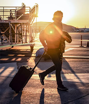 A person rolls a carry-on suitcase beside them on the airport tarmac as a bright sun sets behind them
